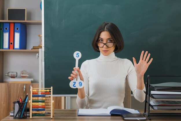 confident young female teacher wearing glasses holding number fan sitting at desk with school tools on in classroom