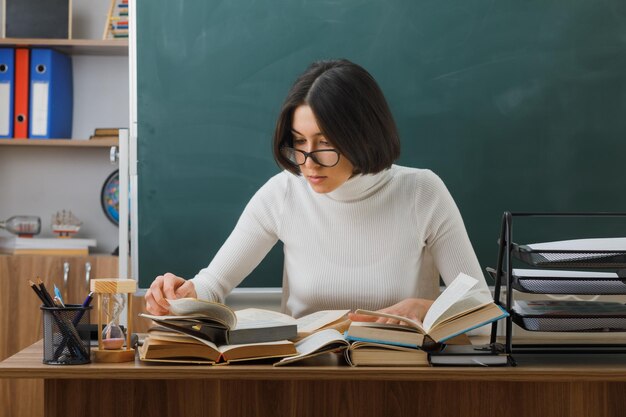 confident young female teacher wearing glasses reading book sitting at desk with school tools on in classroom