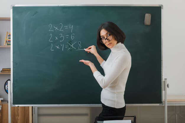 confident young female teacher wearing glasses standing in front blackboard and writes in classroom
