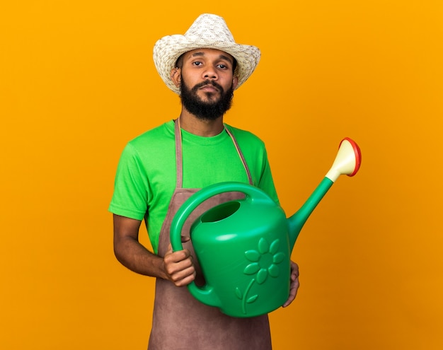 Fiducioso giovane giardiniere afro-americano che indossa un cappello da giardinaggio che tiene in mano un annaffiatoio isolato su un muro arancione