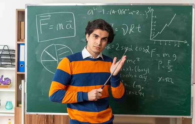 confident young geometry teacher standing in front of chalkboard in classroom holding pointer stick touching hand with it looking at front