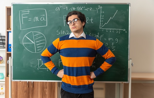 уверенный в себе молодой учитель геометрии в очках, стоящий перед классной доской в классе, держа руки на талии, глядя вперед