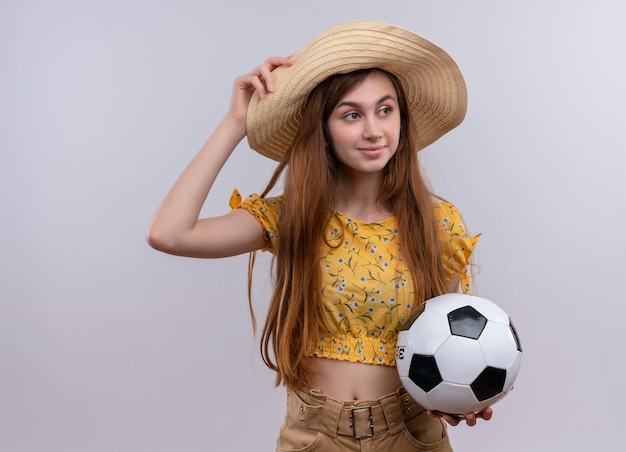 Confident young girl wearing hat holding soccer ball and putting hand on hat on isolated white space