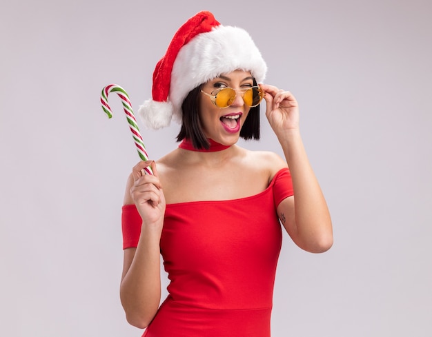 Free photo confident young girl wearing santa hat and glasses holding christmas candy cane looking at camera grabbing glasses winking isolated on white background