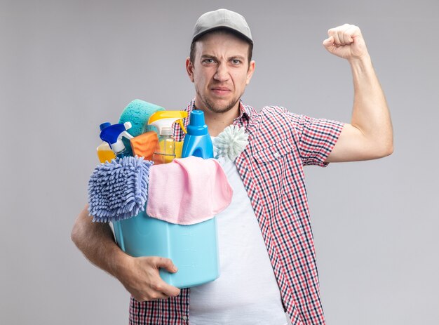 Уверенный молодой парень уборщик в кепке держит ведро с чистящими инструментами, показывая сильный жест, изолированные на белом фоне