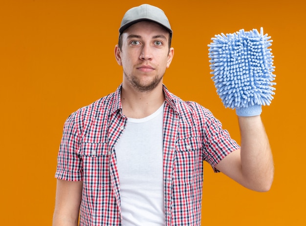 Уверенный молодой парень уборщик в кепке держит тряпку для уборки, изолированную на оранжевой стене