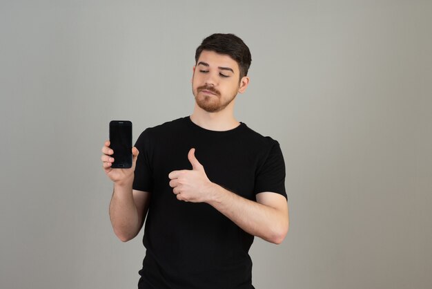 A confident young guy holding a phone and gesturing thumbs up on a grey.