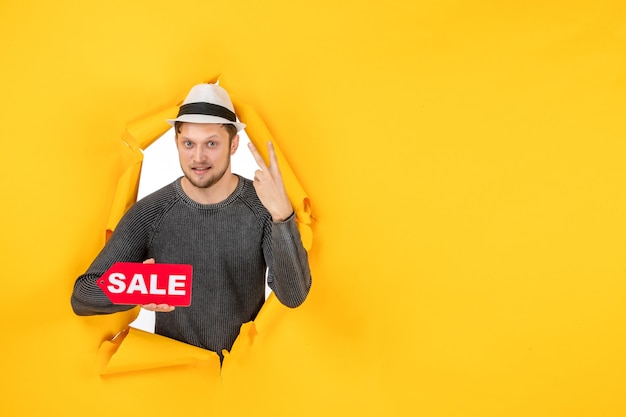 Free photo confident young guy holding sale sign and making victory gesture in a torn on yellow wall