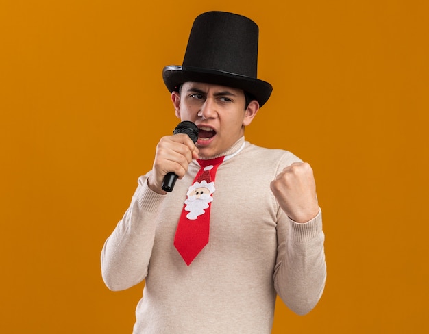 Уверенный молодой парень в шляпе с рождественским галстуком говорит в микрофон, показывая жест да, изолированный на желтой стене