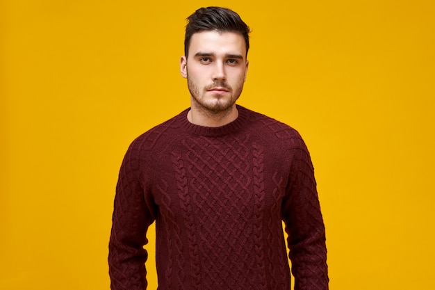 Confident young guy with stubble and dark hair with serious facial expression wearing warm knitted jumper. Brunette man posing  in cozy burgundy sweater, having strict look