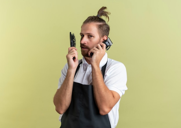Уверенный молодой красивый парикмахер, держащий расческу и машинки для стрижки волос, притворяется, что разговаривает по телефону, используя машинку для стрижки волос в качестве телефона, изолированного на оливково-зеленом с копией пространства