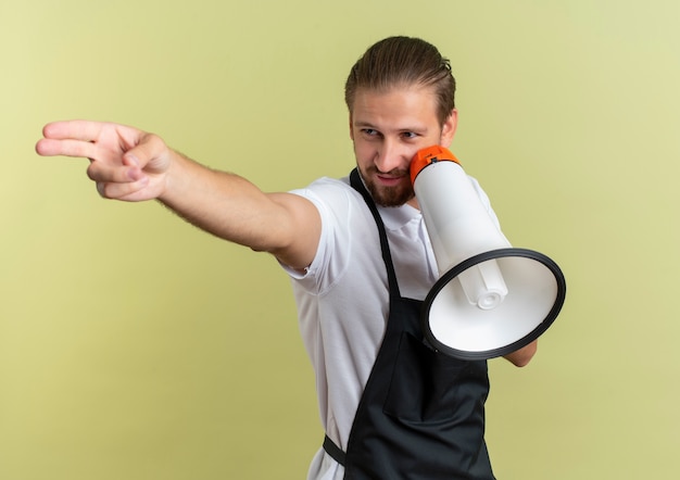 Confident young handsome barber holding speaker looking and pointing at side isolated on olive green