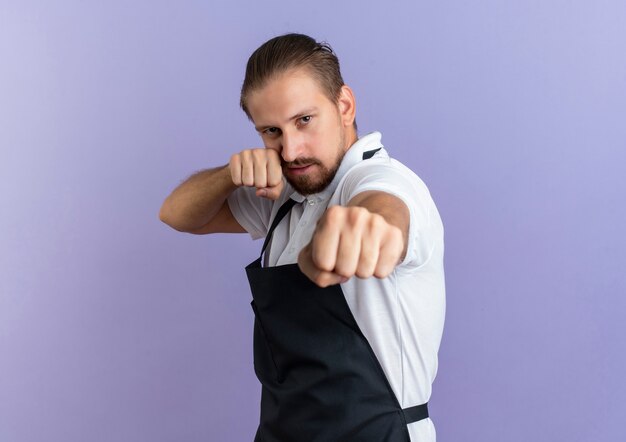 Confident young handsome barber wearing uniform doing boxing gesture isolated on purple  with copy space