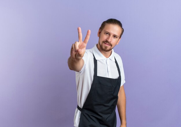 Confident young handsome barber wearing uniform doing peace sign isolated on purple  with copy space
