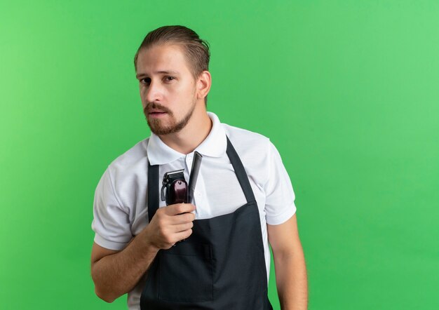 Уверенный молодой красивый парикмахер в униформе, держащей расческу и машинки для стрижки волос, изолированные на зеленом фоне с копией пространства