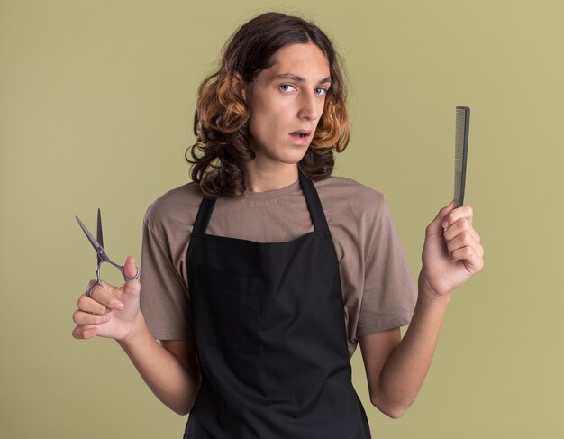 Confident young handsome barber wearing uniform holding comb and scissors