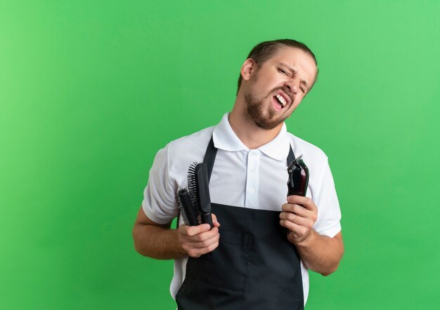 Confident young handsome barber wearing uniform holding combs and hair clippers isolated on green  with copy space