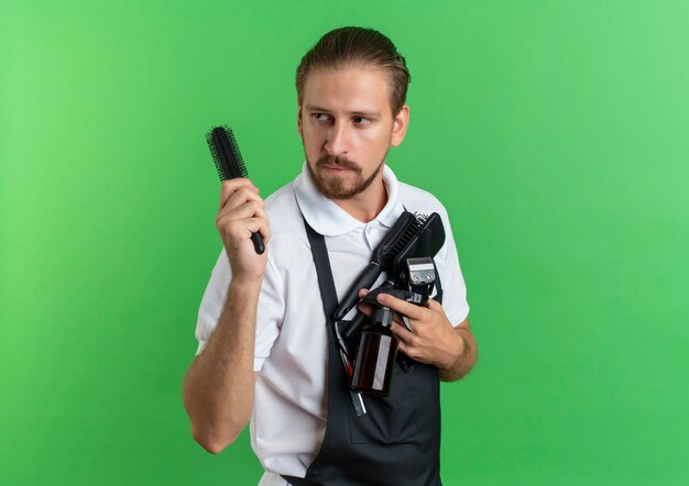 Confident young handsome barber wearing uniform holding combs, spray bottle, hair clippers looking at side isolated on green  with copy space