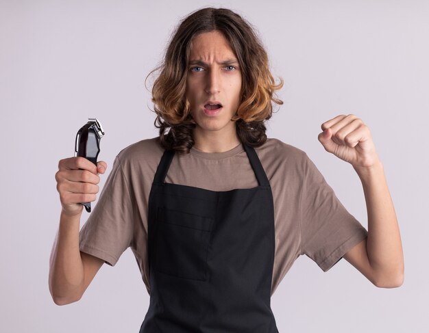 Confident young handsome barber wearing uniform holding hair clippers doing be strong gesture