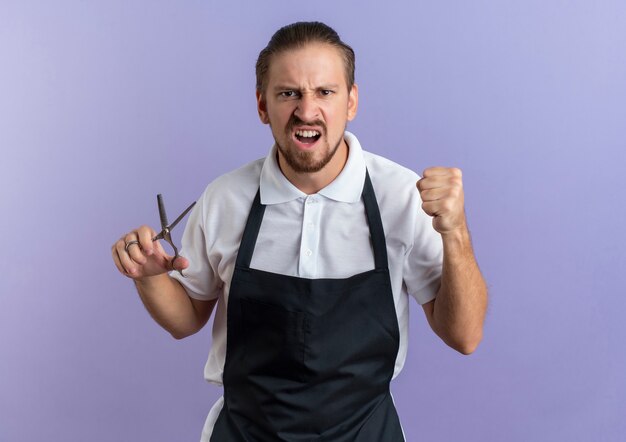 Confident young handsome barber wearing uniform holding scissors and clenching fist isolated on purple  with copy space