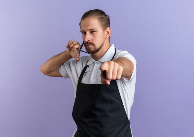 Уверенный молодой красивый парикмахер в униформе, держащий ножницы возле лица и указывающий на фиолетовый с копией пространства
