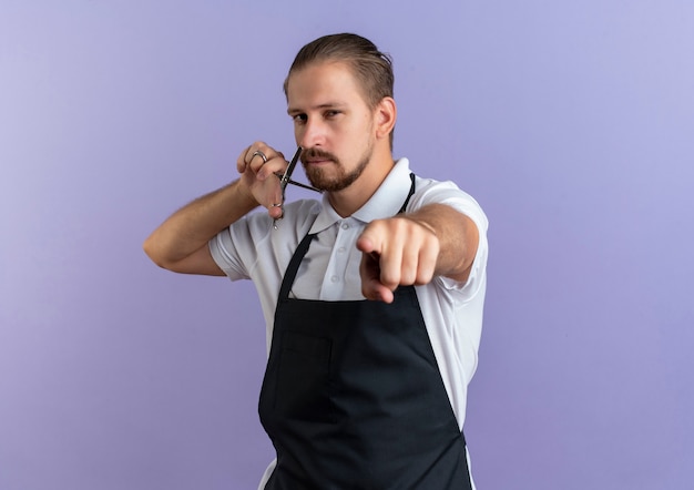 Confident young handsome barber wearing uniform holding scissors near face and pointing isolated on purple  with copy space