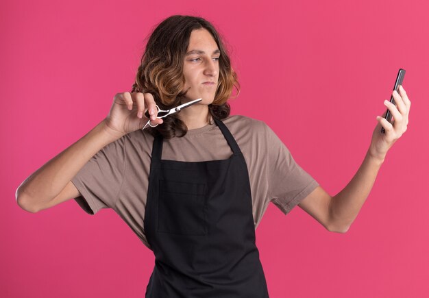 Confident young handsome barber wearing uniform holding scissors taking selfie 