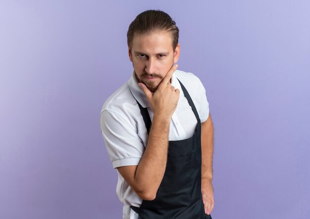 Confident young handsome barber wearing uniform putting hand on chin and another one on waist isolated on purple  with copy space