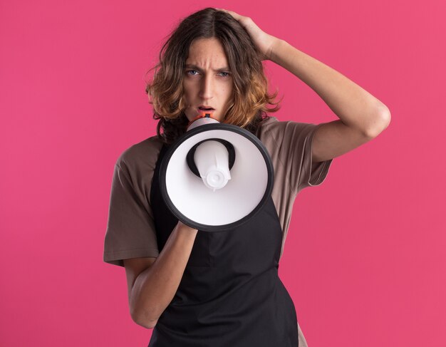 Confident young handsome barber wearing uniform putting hand on head  talking by speaker isolated on pink wall