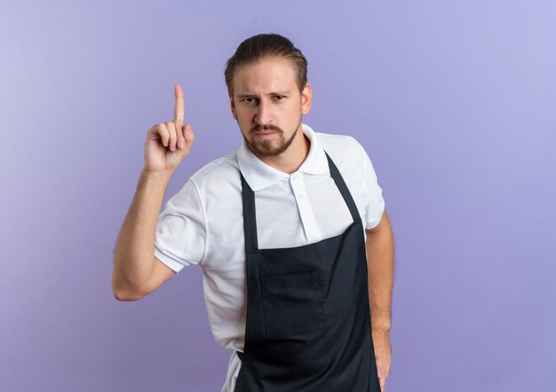 Free photo confident young handsome barber wearing uniform putting hand on waist and pointing up isolated on purple  with copy space