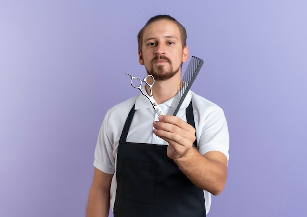 Confident young handsome barber wearing uniform stretching out comb and scissors towards camera isolated on purple  with copy space