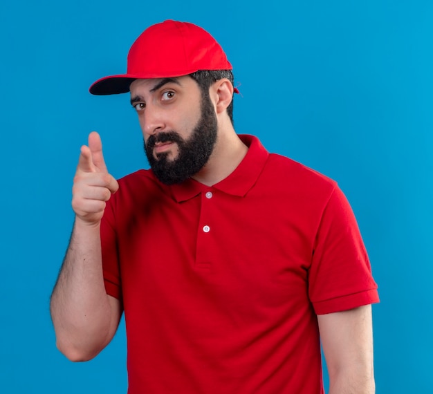 Confident young handsome caucasian delivery man wearing red uniform and cap looking and pointing isolated on blue 