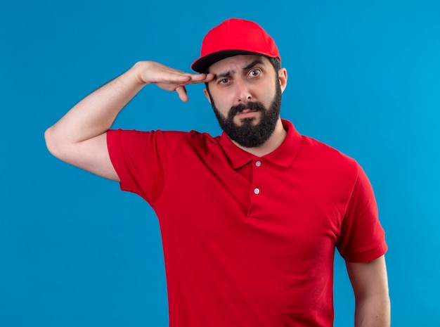 Confident young handsome caucasian delivery man wearing red uniform and cap putting hand near head isolated on blue 
