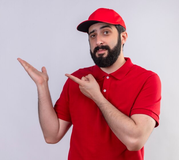 Confident young handsome caucasian delivery man wearing red uniform and cap showing empty hand and pointing at it isolated on white 