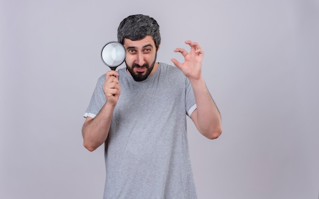 Confident young handsome caucasian man holding magnifying glass and gesturing tiger paw isolated on white  with copy space