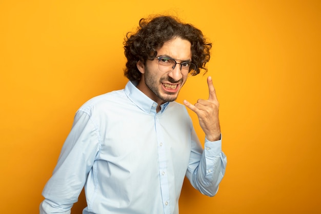 Confident young handsome caucasian man wearing glasses looking at camera showing teeth doing rock sign isolated on orange background with copy space