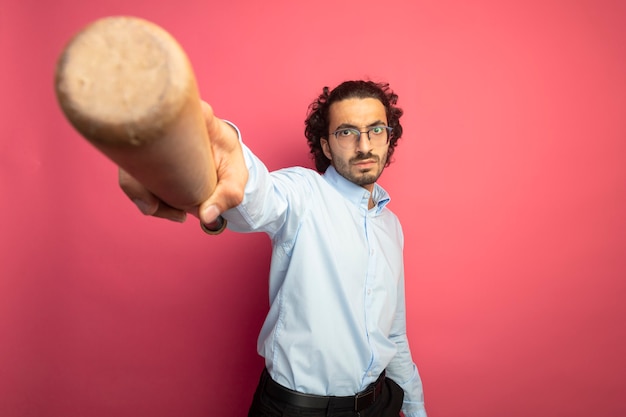 Fiducioso giovane uomo caucasico bello con gli occhiali che guarda l'obbiettivo allungando la mazza da baseball verso la telecamera isolata su sfondo cremisi con spazio di copia