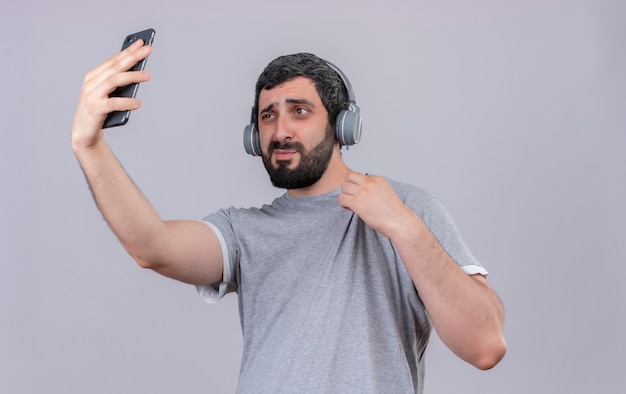 Confident young handsome caucasian man wearing headphones holding his shirt and taking selfie isolated on white  with copy space