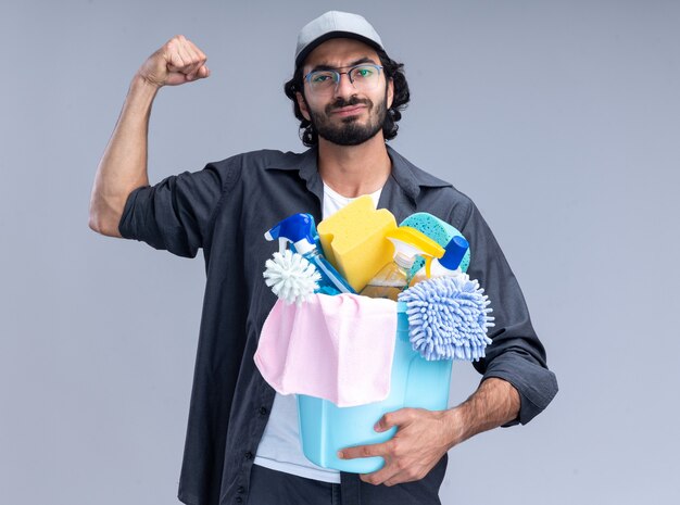 Уверенный молодой красивый парень-уборщик в футболке и кепке, держащий ведро с чистящими средствами, демонстрирует сильный жест, изолированный на белой стене