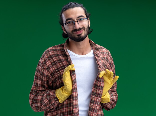 Уверенный молодой красивый парень-уборщик в футболке и перчатках держит футболку, изолированную на зеленой стене