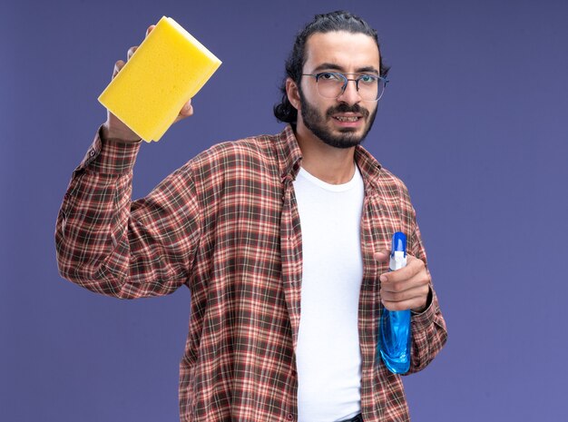 Confident young handsome cleaning guy wearing t-shirt holding spray bottle with sponge isolated on blue wall