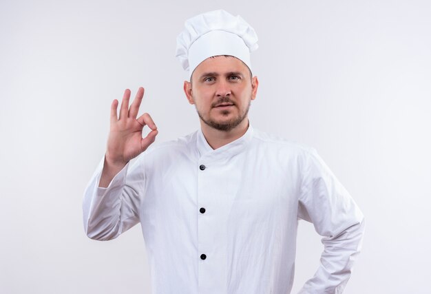 Confident young handsome cook in chef uniform doing ok sign isolated on white wall