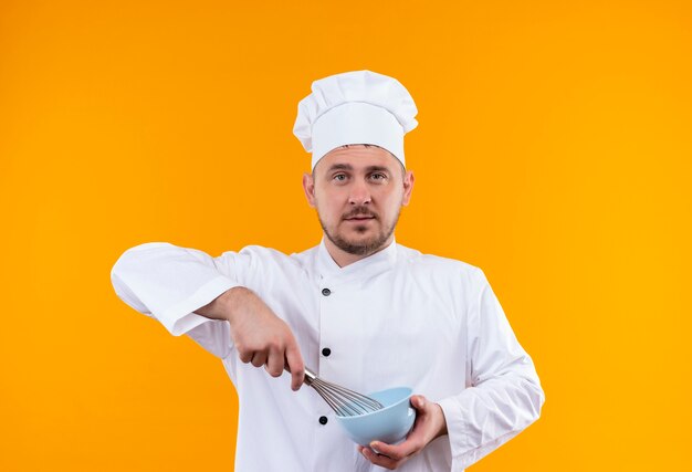 Confident young handsome cook in chef uniform holding bowl and whisk isolated on orange wall