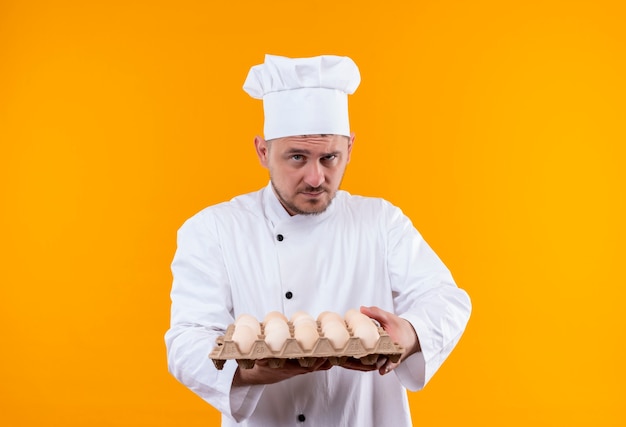 Confident young handsome cook in chef uniform holding carton of eggs isolated on orange wall