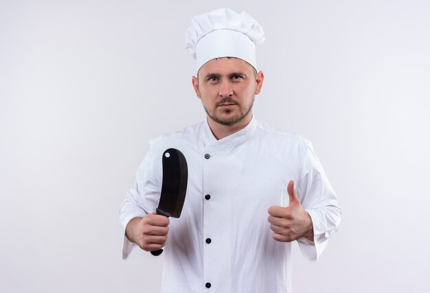 Confident young handsome cook in chef uniform holding cleaver and showing thumb up on isolated white wall