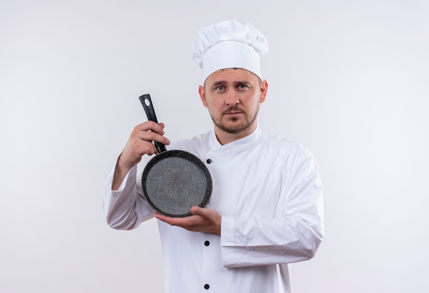 Confident young handsome cook in chef uniform holding frying pan isolated on white wall