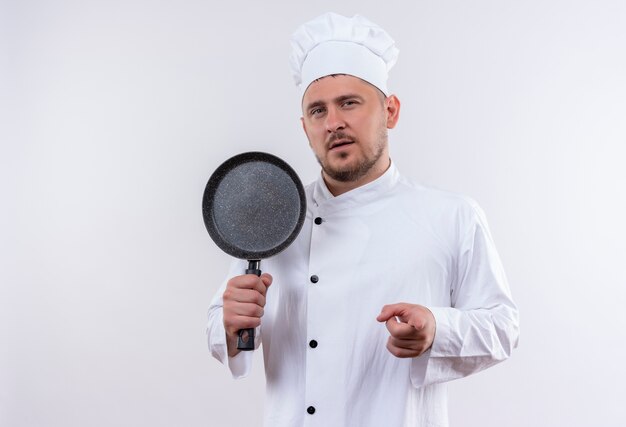 Confident young handsome cook in chef uniform holding frying pan and pointing on isolated white wall with copy space