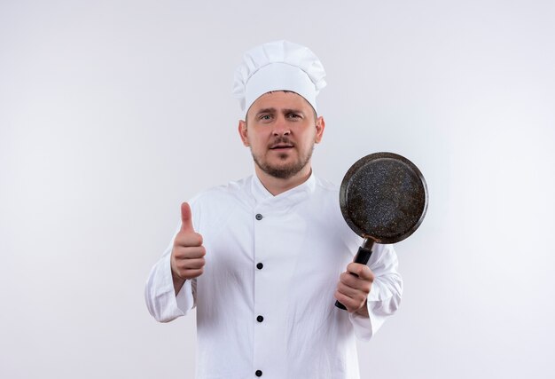 白い壁に孤立した親指を現してフライパンを保持しているシェフの制服を着た自信のある若いハンサムな料理人