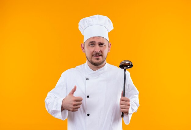 Confident young handsome cook in chef uniform holding ladle and showing thumb up on isolated orange wall with copy space