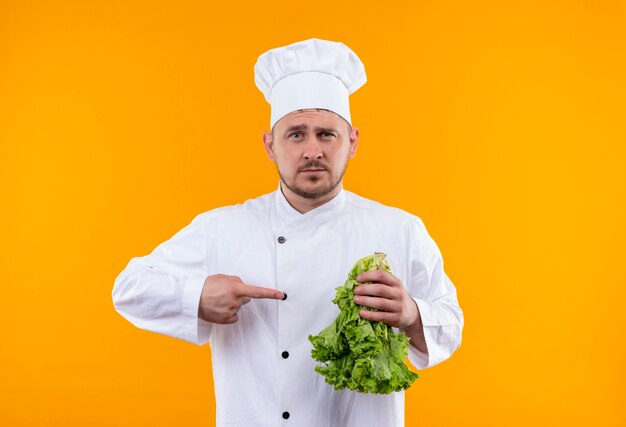 Уверенный молодой красивый повар в униформе шеф-повара держит салат и указывает на него, изолированном на оранжевой стене